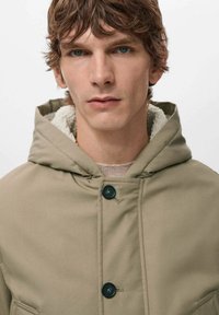 Chaqueta con capucha color caqui y un suave forro crema, con dos botones negros y un diseño simple y funcional. Tela suave con un corte holgado.