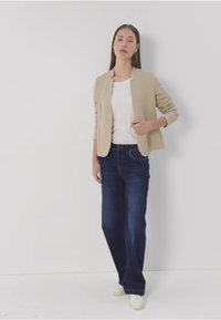 Beige blejzr přes bílé tričko, spárovaný s tmavě modrými džíny a světlými teniskami. Blejzr má úzký střih a mírně texturovanou tkaninu.