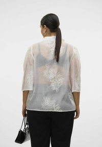 Eine Frau mit dunklem Haar im Pferdeschwanz trägt eine transparente weiße Bluse mit Blumenmuster über einem grauen Tanktop, schwarze Hose und hält eine schwarze Handtasche.