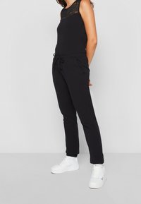 Kvinna i ärmlös svart spetsblus, svarta dragsnoddbyxor och vita sneakrar som står mot en ljus, enfärgad bakgrund.