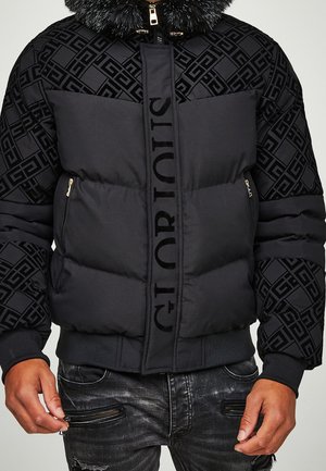Sort dunsjakke med pelskrave, geometriske mønstre på ærmerne og teksten "G TOTR OUIS" på frontlynlåsdækslet, båret med sorte slidte jeans.