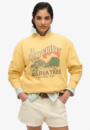 Sweatshirt en polaire jaune avec un design graphique portant "SuperDry" et "Joshua Tree National Park". Associé avec un short clair et une chemise rayée en dessous.