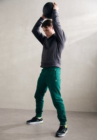 Pantaloni della tuta verdi con cerniere laterali, abbinati a un hoodie grigio. Il modello indossa sneakers nere con dettagli neon, tenendo un pallone da basket sopra la testa.