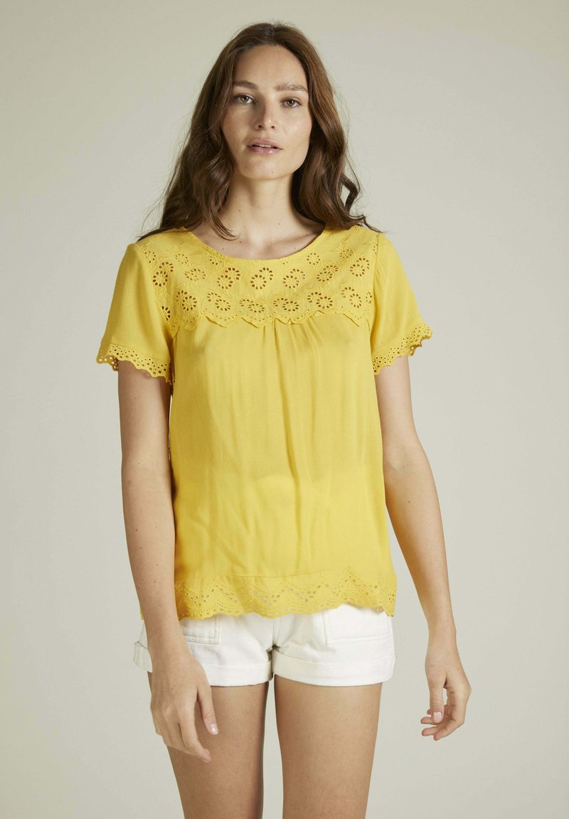Blouse jaune à manches courtes, présentant des détails en œillets floraux le long de l'encolure et de l'ourlet. Texture lisse, coupe ample, associée à un short blanc.
