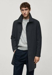 Mørk grå uldfrakke med høj krave og frontlukning med knapper, båret over en lys grå sweater og mørke bukser. Enkel design, ingen mønstre.