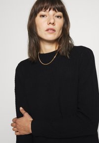 Zwart geribbeld sweater met een ronde halslijn en lange mouwen, gekenmerkt door een gestructureerde stof en een getailleerde pasvorm, gecombineerd met een gouden ketting.}}