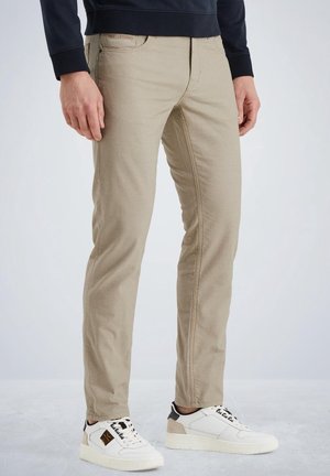 Mann trägt beige Hose und weiße Turnschuhe, steht vor einem einfarbigen hellen Hintergrund.