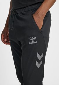 Schwarze Sporthose aus glattem Stoff mit Kordelzug am Bund, Seitentaschen sowie grauem Logo und Chevron-Akzenten am Bein.