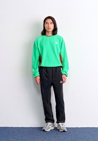 Zeleno flis jopico z rjavimi detajli in majhnim belim logotipom, v kombinaciji z črnimi trenirkami in sivimi športnimi čevlji. Enostavna ozadje.