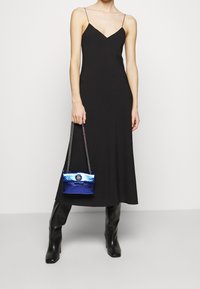 Abito nero lungo fino alle caviglie con spalline sottili, abbinato a una piccola borsa a tracolla in pelle di serpente blu con logo e tracolla a catena. Stivali neri fino al ginocchio.