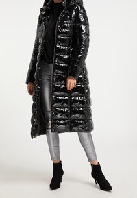 Černá lesklá péřová bunda s kapucí, vybavená zipem a druky. Nošena přes lesklé stříbrné legíny a černé kotníkové boty.