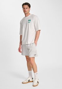White cotton t-shirt with a green graphic logo, paired with gray shorts and white sneakers featuring black accents and a gum sole.