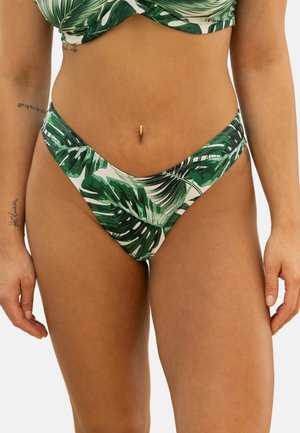 Frau trägt grünen und weißen Bikini mit tropischem Blattmuster, sichtbare Tattoos an den Armen und einen Bauchnabelpiercing.