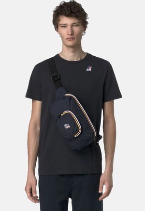 Giovane uomo che indossa una maglietta K-Way nera e una borsa a tracolla blu navy con cerniere a strisce su sfondo chiaro.