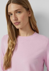 Różowy sweter z okrągłym dekoltem, gładką teksturą i długimi rękawami, prezentowany na modelce o prostych włosach. Minimalistyczny design.