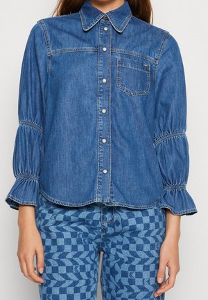 Kvinde iført en blå denimskjorte med knapper med puffede, flæseærmer og ternede blå denimbukser.