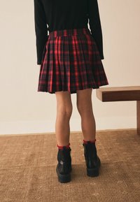Červená kostkovaná plisovaná sukně s vysokým pasem, doplněná černými botami se červenými volánkovými akcenty nahoře.