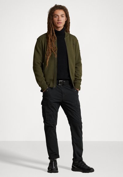 Casaco bomber verde-oliva por cima de um suéter de gola alta preto, combinado com calças cargo pretas e ténis pretos. Apresenta detalhes de fecho éclair e bolsos.