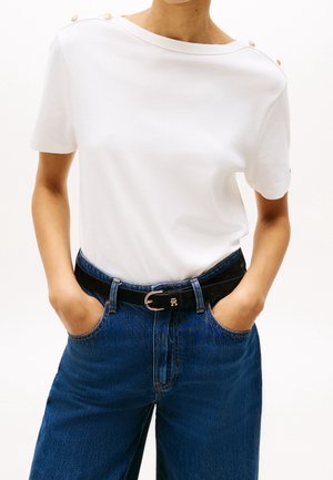 Top blanco de manga corta con escote redondo y detalles de botones dorados en el hombro, combinado con jeans azul de cintura alta y un cinturón negro.