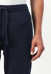 Mørkeblå sweatpants med elastisk talje, snøre, sidelomme med lynlås og en glat bomuldsstruktur. Hånd hviler på stoffet.