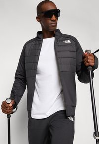 Schwarze Zip-Jacke mit gepolstertem Vorderteil, grauen Ärmeln und Logo. Darunter ein weißes T-Shirt, kombiniert mit dunklen Hosen. Der Benutzer hält Trekkingstöcke.