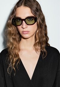 Femme aux longs cheveux ondulés portant des lunettes de soleil rectangulaires vert foncé et un top noir à col en V, sur un fond léger uni.