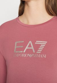 Långärmad t-shirt i mjuk rosa tyg med en metallisk silverlogotyp med texten "EA7 EMPORIO ARMANI" och randiga detaljer.