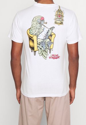 Bílé tričko s podrobnou grafikou zeleného papouška sedícího na žluté křeslo, který drží dýmku, s ptačí klecí nad hlavou.