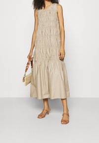 Robe sans manches beige avec texture froncée, bas évasé et coupe loose. Associée à des sandales à lanières marron et un petit sac à main en osier.