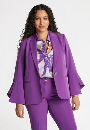 Blazer viola con maniche a campana, tessuto morbido, chiusura a un bottone e tasche frontali, indossato sopra una blusa a fantasia con sfumature di blu e rosa.