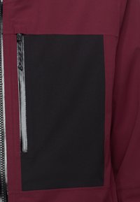 Maroon jacka med en stor svart panel, som har en synlig dragkedjeficka. Tyget verkar slätt med subtila sömdetaljer.