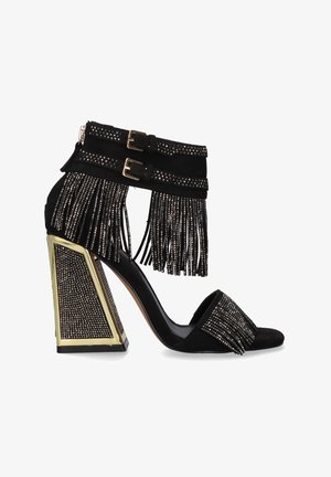 Sandalia negra de tacón alto con tacón bloque adornado con detalles dorados, doble hebilla en las correas del tobillo y detalles de flecos brillantes en las correas y en la puntera.