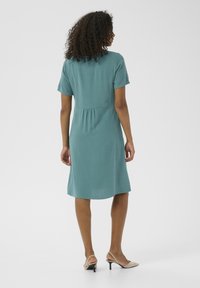 Ein türkisfarbenes Kurzarmkleid mit einem taillierten Oberteil, geraffter Taille und fließendem Rock. Getragen mit beigen hohen Sandalen.