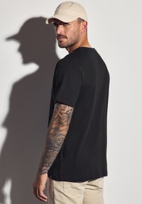 Černé tričko s krátkým rukávem, vyrobené z bavlny, nošeno mužem v béžové kšiltovce. Viditelné tetování na rukávech a světlé kalhoty.