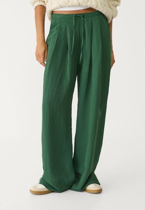 Femme portant un pantalon vert ample taille haute avec un lien devant, associé à des baskets blanches et un pull en maille crème.