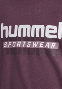 Dunkelrotes T-Shirt aus Baumwollmischung mit großem weißem "hummel"-Logo und "SPORTSWEAR"-Schriftzug, versehen mit einem strukturierten Hintergrundmuster.