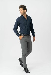 Mand går med hænderne delvist i lommerne, iført en mørkeblå skjorte, grå bukser, sort bælte og sorte dress sko på en hvid baggrund.