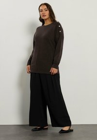Maglione marrone a maglia con scollo rotondo, caratterizzato da tre bottoni metallici sulla spalla sinistra. Abbinato a pantaloni larghi neri e scarpe nere.