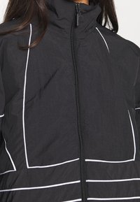 Svart jacka med dragkedja fram, hög krage och vita horisontella ränder. Tillverkad av lätt, strukturerat material.