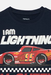 Mørk marineblå T-skjorte med et motiv av en rød racerbiler med nummer 95, teksten "I AM LIGHTNING", og en svart-hvit rutete ramme.