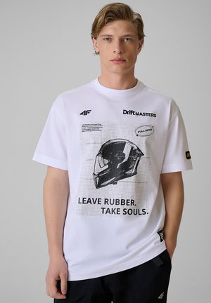 Młody mężczyzna o blond włosach, ubrany w białą koszulkę z grafiką hełmu i napisem "Leave Rubber. Take Souls." oraz czarne spodnie.
