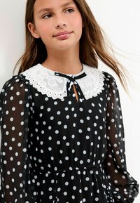 Junges Mädchen mit langen Haaren, das ein schwarzes durchscheinendes Kleid mit Punkten und einem weißen Spitzenkragen sowie einer kleinen schwarzen Fliege trägt.