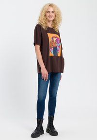 Brązowy T-shirt z kolorowym wzorem graficznym przedstawiającym osobę, w połączeniu z niebieskimi skinny jeansami i czarnymi sznurowanymi botkami.