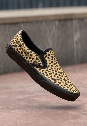Slip-on shoe with yellow and black spotted pattern and black sole, floating above a pinkish concrete surface.