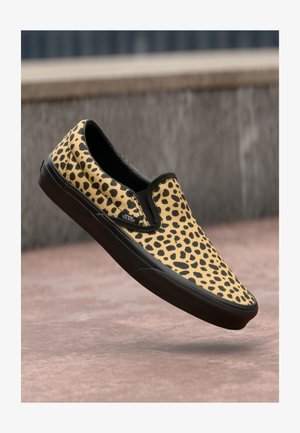 Slip-On-Schuhe mit gelb-schwarzem gepunktetem Muster und schwarzer Sohle, schwebend über einer rosa betonten Betonfläche.