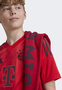 Maillot de sport rouge avec un col en V, logo du FC Bayern München et logo du sponsor, associé à une écharpe rouge avec des lettres et motifs foncés.