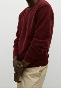 Maglione bordeaux a maglia con scollatura rotonda e polsini a coste, abbinato a pantaloni di colore chiaro. Il maglione ha una finitura morbida e texturizzata.