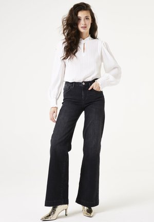 Weiße, texturierte Bluse mit Schlitzdetail, kombiniert mit hoch taillierten, dunklen Jeans im ausgestellten Schnitt und metallisch goldenen spitzen High Heels.