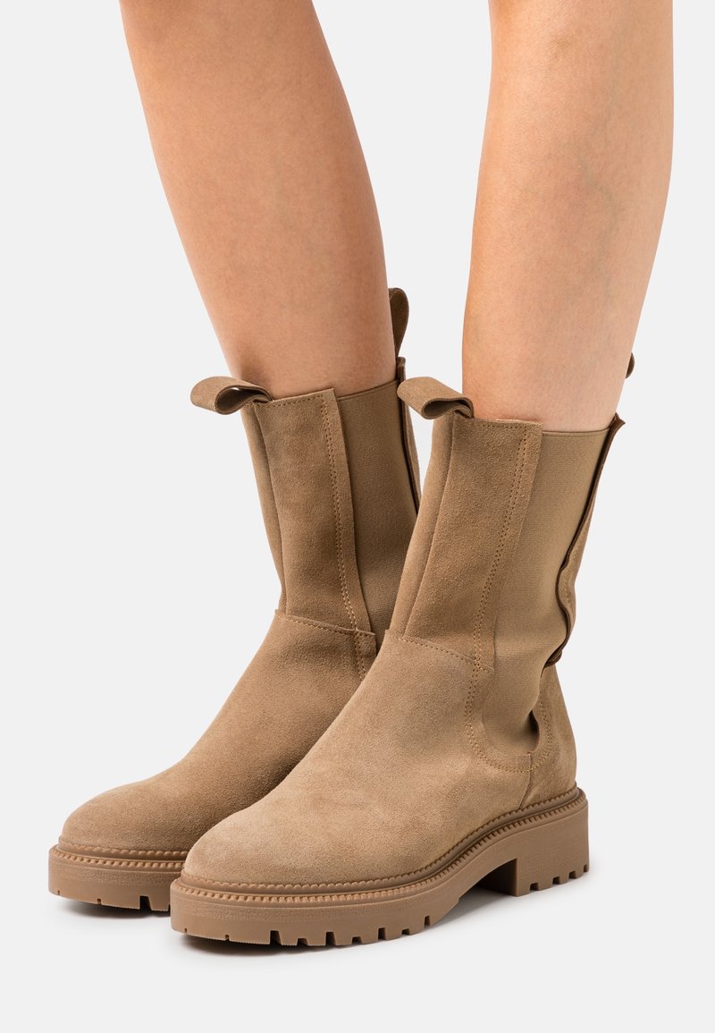 Bottes mi-mollet en daim marron clair avec languettes de tirage, panneaux latéraux élastiques et semelles crantées, portées sur des jambes nues.