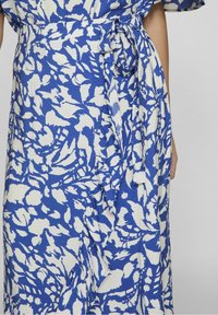 Modrá a bílá květinová zavinovací šaty vyrobená z texturované tkaniny s opaskem na pase. Má volné rukávy a délku ke kolenům.